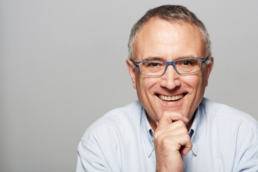 senior man wearing glasses smiling and showing off implant-supported dentures in front of gray background