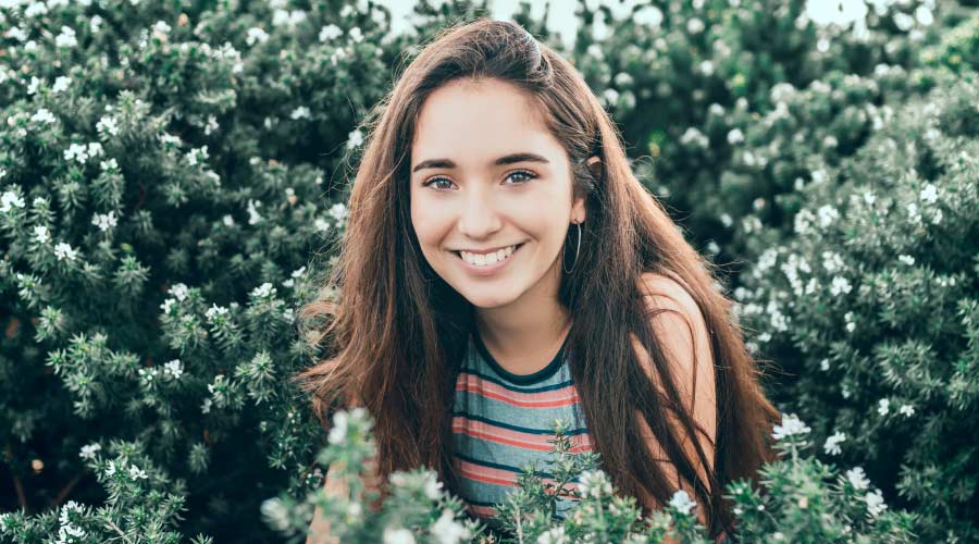 Brunette young woman smiles while standing in a cluster of green bushes with white flowers and piney needles