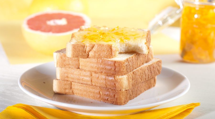 A stack of white bread with the top piece smothered in orange marmalade on a white plate in front of half a grapefruit