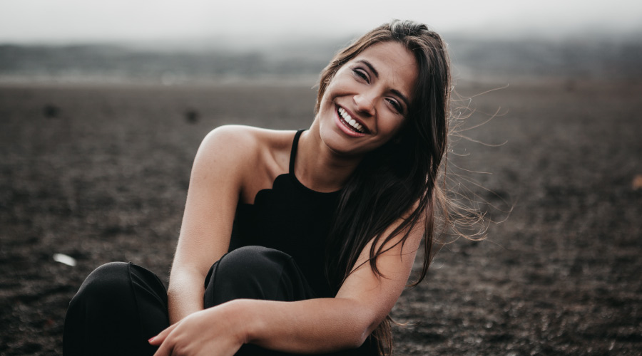 Brunette woman smiles with dental implants while wearing a black tanktop and sitting in a dirt field