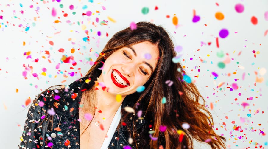 A brunette woman wearing red lipstick smiles as colorful confetti falls celebrating the move of the dental office