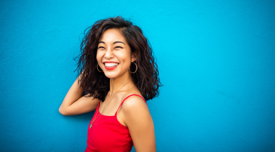 Brunette woman in a red tanktop smiles against a blue wall because she is pain-free after root canal therapy