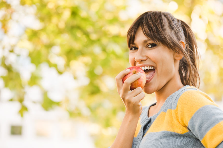 Girl eating an apple outdoors in front of a leafy background. 